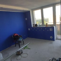 MagMarBud - Świeżo pomalowane na niebiesko ściany w pokoju z widocznymi narzędziami malarskimi, otwartymi drzwiami balkonowymi i widokiem na zewnątrz.