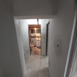 MagMarBud - Widok korytarza z wejściem do łazienki w trakcie prac wykończeniowych; ściany i podłoga w szarym betonie, widoczne ślady po pracach, w łazience częściowo zamontowana armatura i meble łazienkowe.