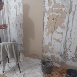MagMarBud - Ściana w trakcie przygotowania do malowania: częściowo otynkowana, częściowo z resztkami starej farby, zabezpieczona podłoga, widoczne narzędzia i wiaderka z zaprawą.