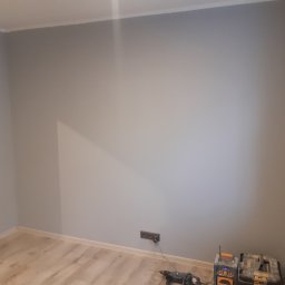 MagMarBud - Świeżo pomalowane na szaro ściany pokoju z widocznymi narzędziami malarskimi i wiertarką przy podłodze, ukazujące etap prac wykończeniowych.