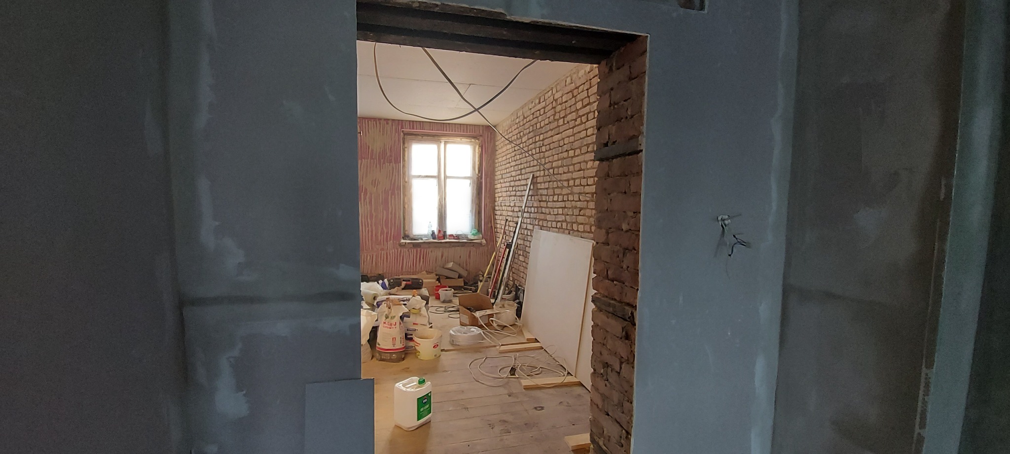 Wnętrze w trakcie prac przygotowawczych do wykończenia: surowa cegła, częściowo otynkowane ściany, widoczne worki z materiałami budowlanymi i narzędzia na drewnianej podłodze, okno z zasłoniętą szybą.