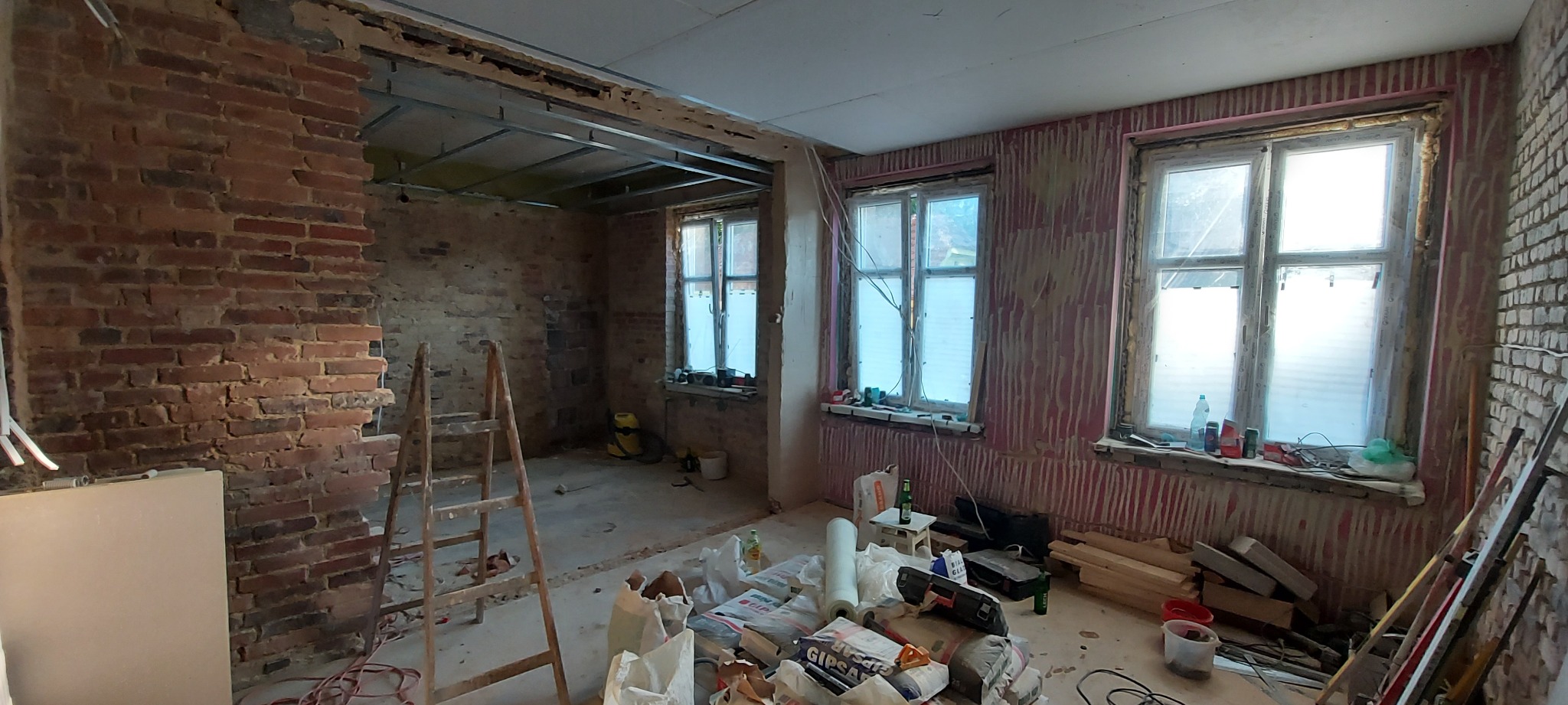 Wnętrze mieszkania w trakcie remontu: odsłonięta cegła na ścianie, częściowo zamontowany sufit podwieszany z metalowych profili, okna z widocznymi śladami prac, worki z gipsem i narzędzia...