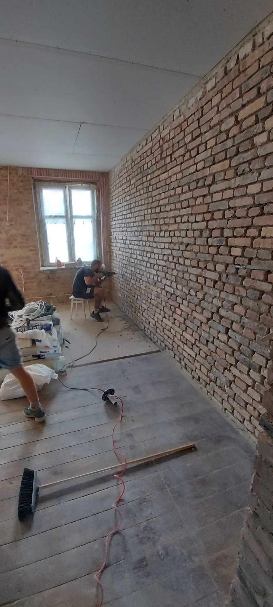 Mężczyzna wierci w ceglanej ścianie w trakcie prac remontowych, widoczne narzędzia i materiały budowlane, podłoga drewniana częściowo zakryta folią ochronną.