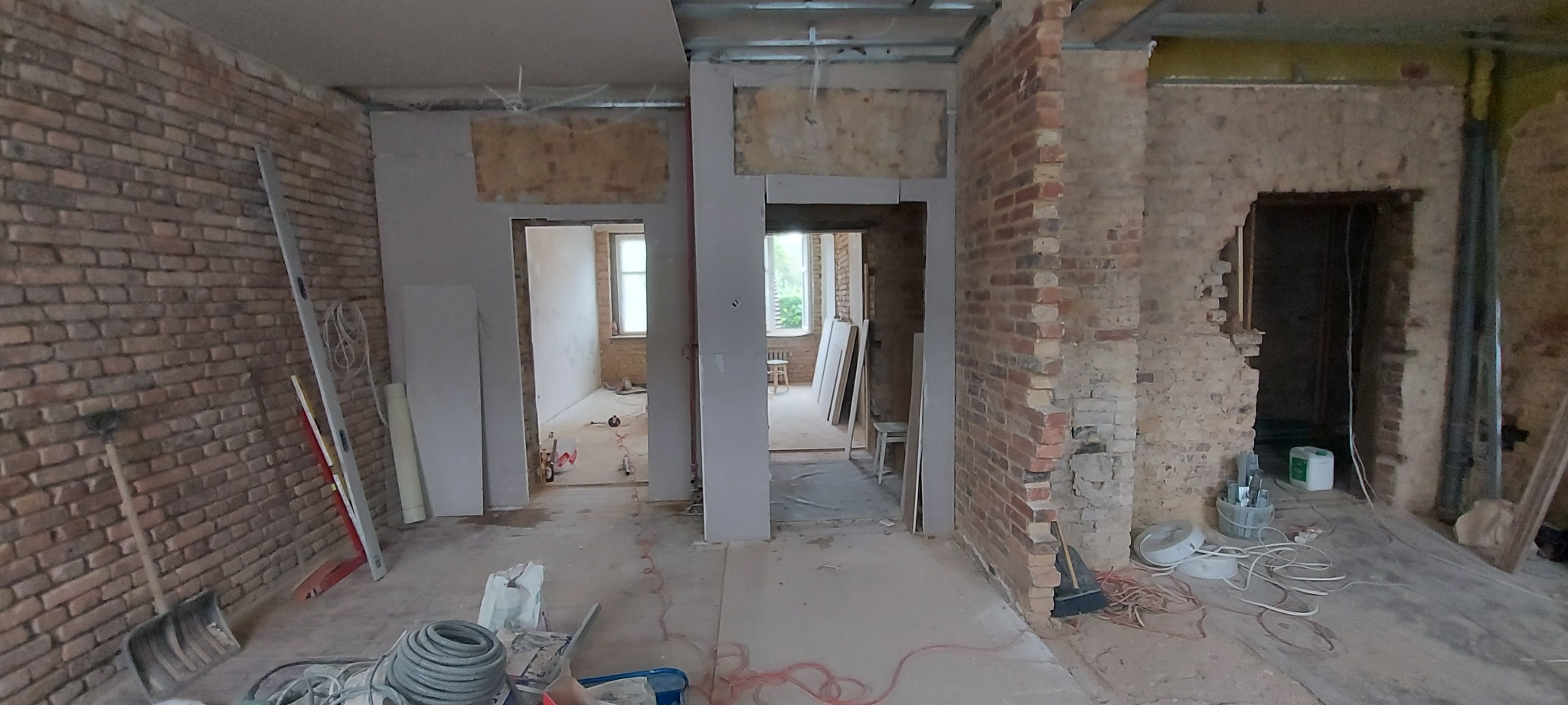 Wnętrze w trakcie prac remontowych: odsłonięta cegła, metalowa konstrukcja sufitu, przygotowane płyty kartonowo-gipsowe, widoczne narzędzia i materiały budowlane, stan surowy z widokiem na kolejne...