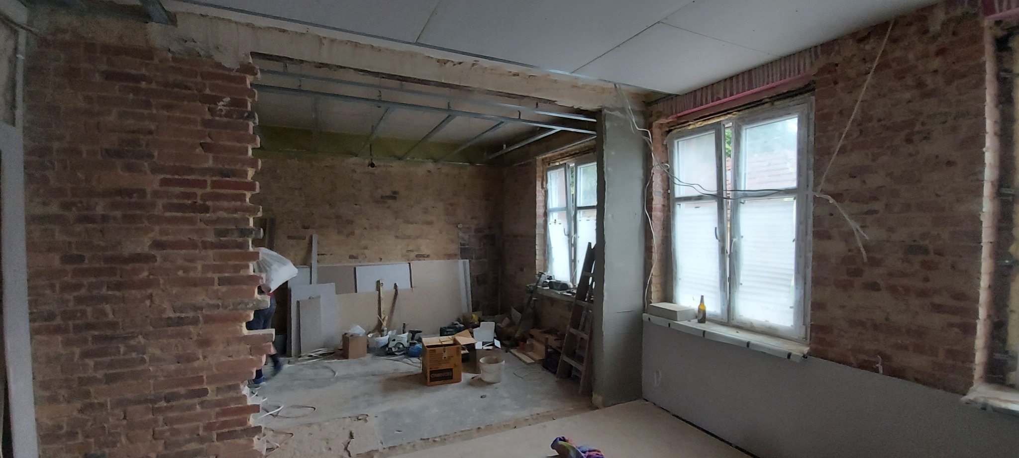 Wnętrze w trakcie remontu: odsłonięta cegła, metalowa konstrukcja sufitu podwieszanego, częściowo położone płyty gipsowo-kartonowe na ścianach, okna z żaluzjami, narzędzia i materiały budowlane...