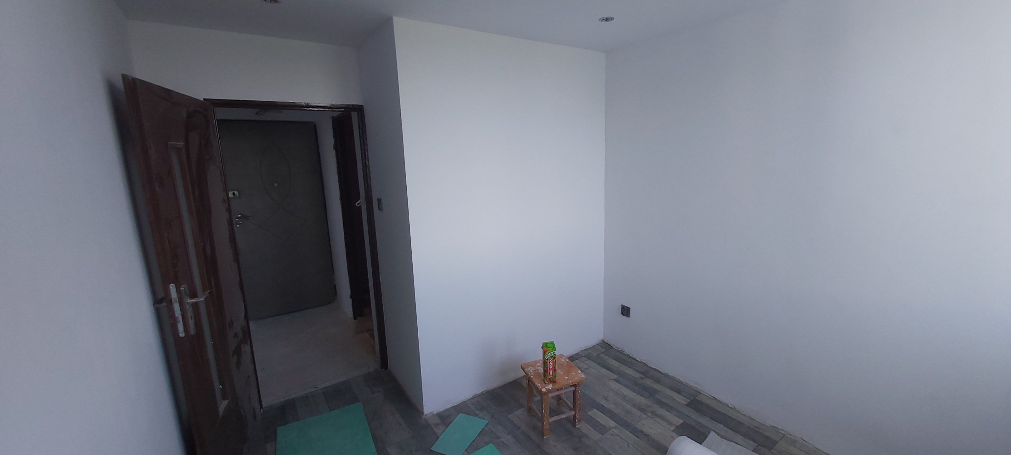 Pokój po świeżym malowaniu, z otwartymi drzwiami, podłogą z paneli i stołkiem z sokiem w rogu, przygotowany do dalszych prac wykończeniowych.