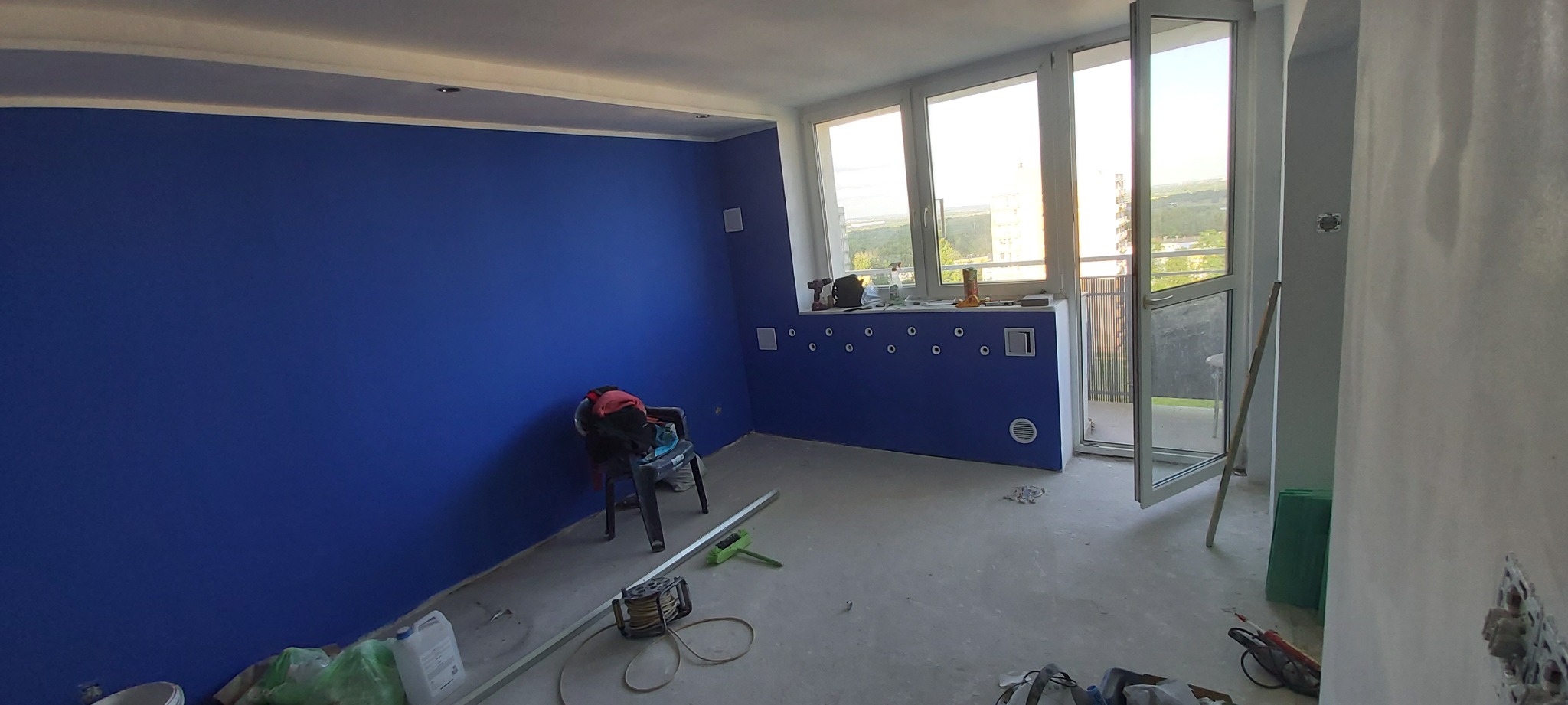 Świeżo pomalowane na niebiesko ściany w pokoju z widocznymi narzędziami malarskimi, otwartymi drzwiami balkonowymi i widokiem na zewnątrz.