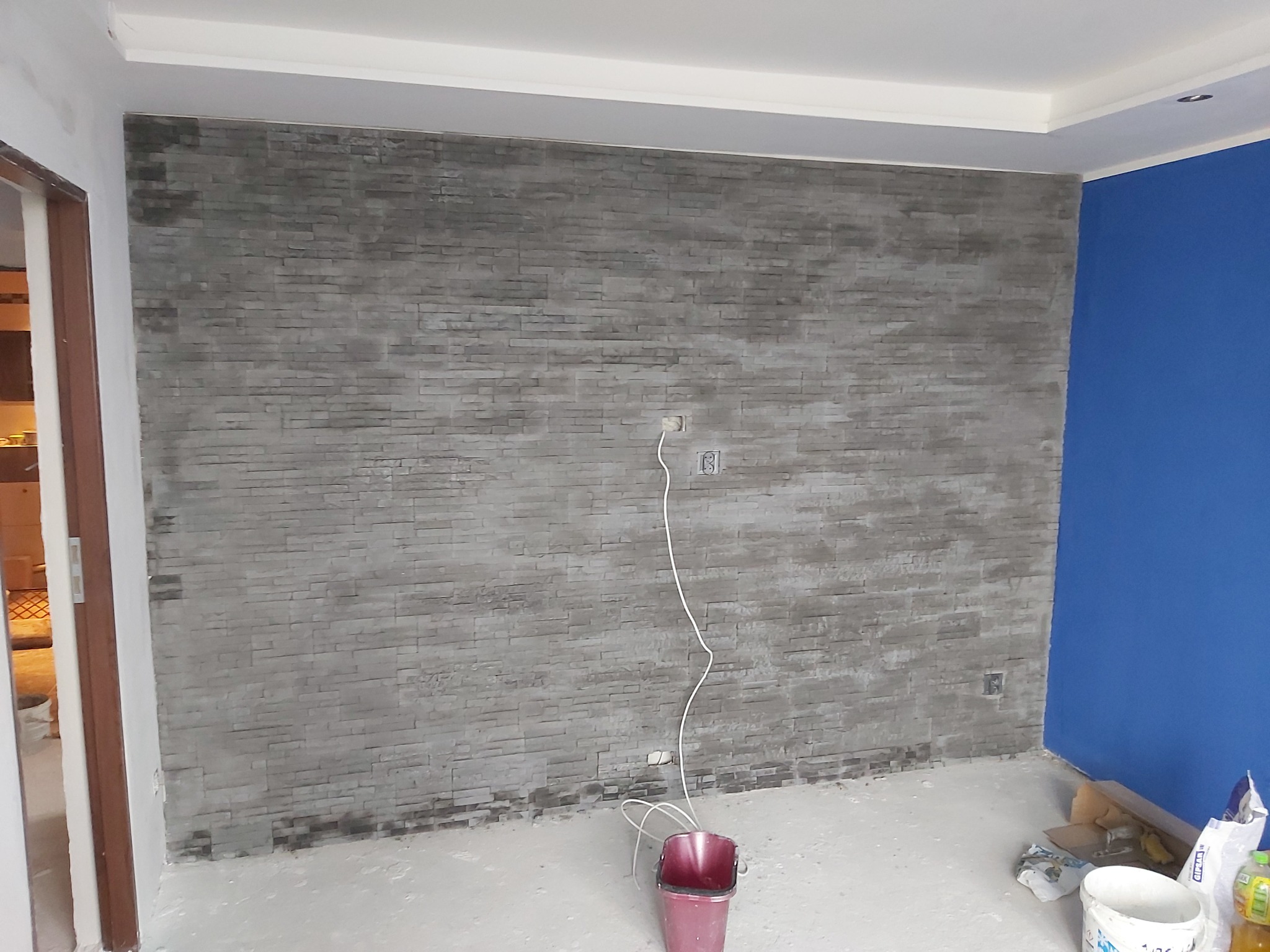 Ściana wykończona szarą cegłą dekoracyjną w trakcie prac remontowych, widoczne puszki elektryczne, kabel, wiadro i materiały budowlane przy podłodze, fragment pomalowanej na niebiesko ściany w rogu.
