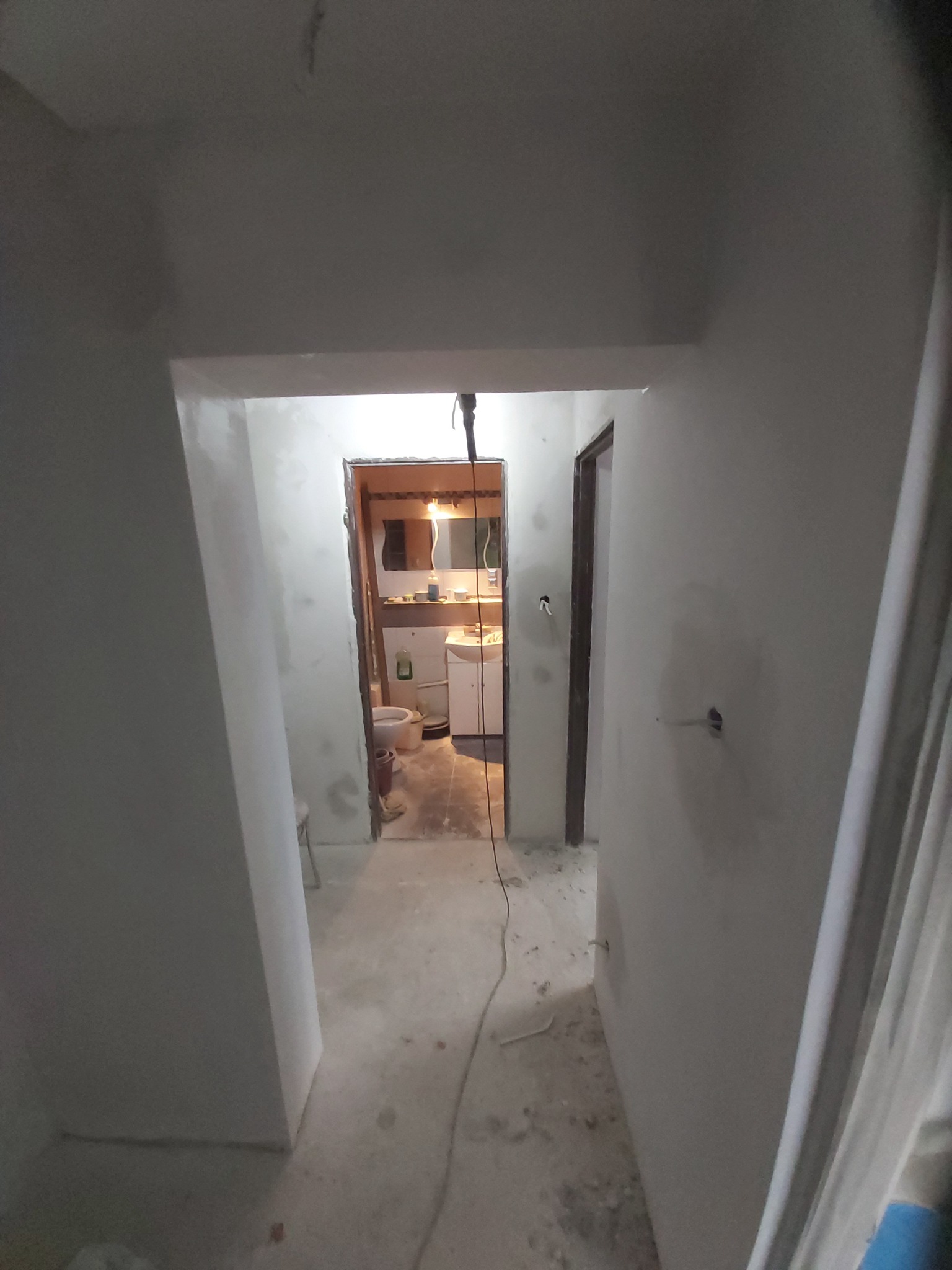 Widok korytarza z wejściem do łazienki w trakcie prac wykończeniowych; ściany i podłoga w szarym betonie, widoczne ślady po pracach, w łazience częściowo zamontowana armatura i meble łazienkowe.