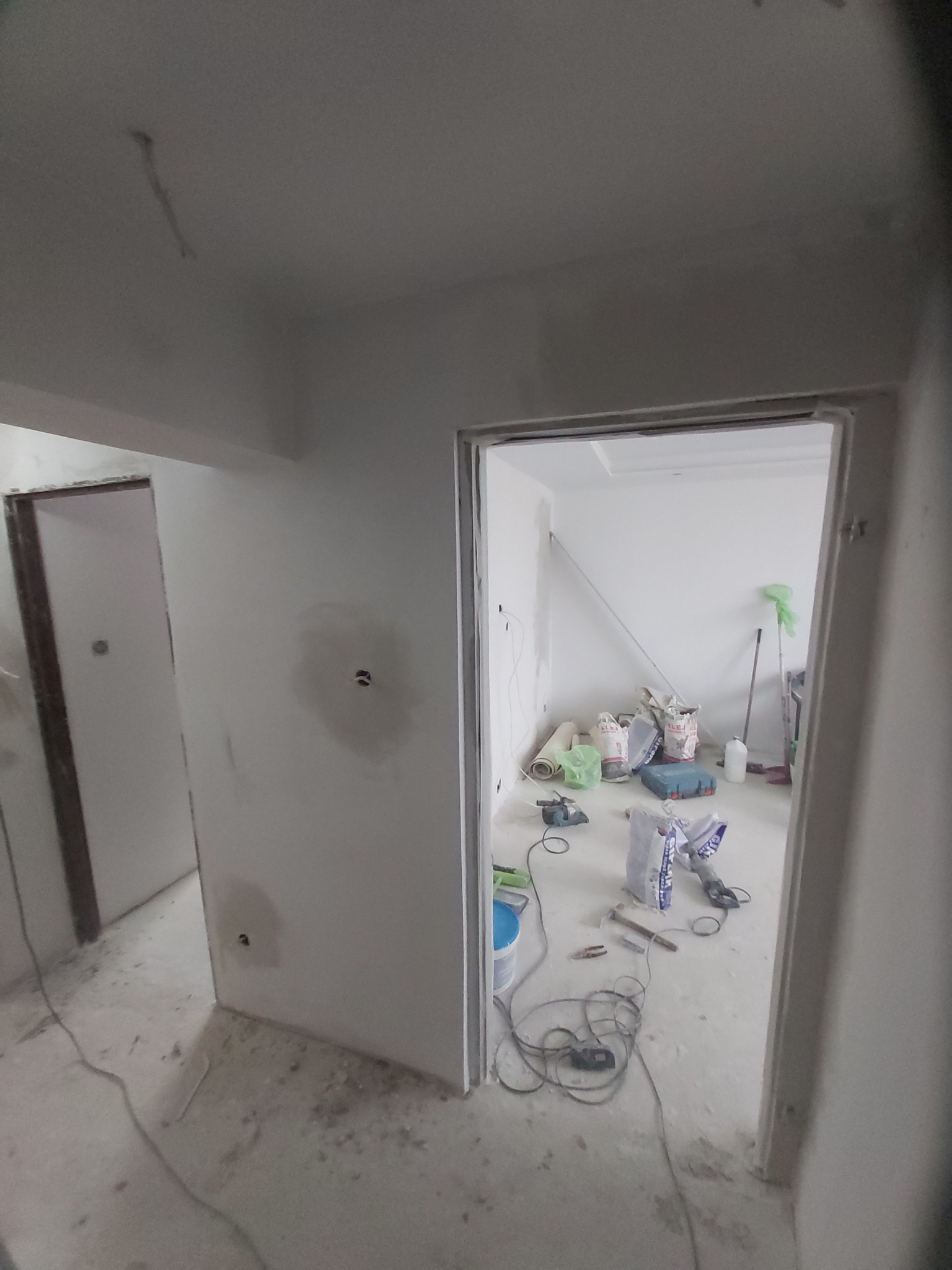 Wnętrze mieszkania w trakcie prac wykończeniowych, widoczne ściany przygotowane do malowania, narzędzia i materiały budowlane na podłodze, widok przez dwa otwory drzwiowe.