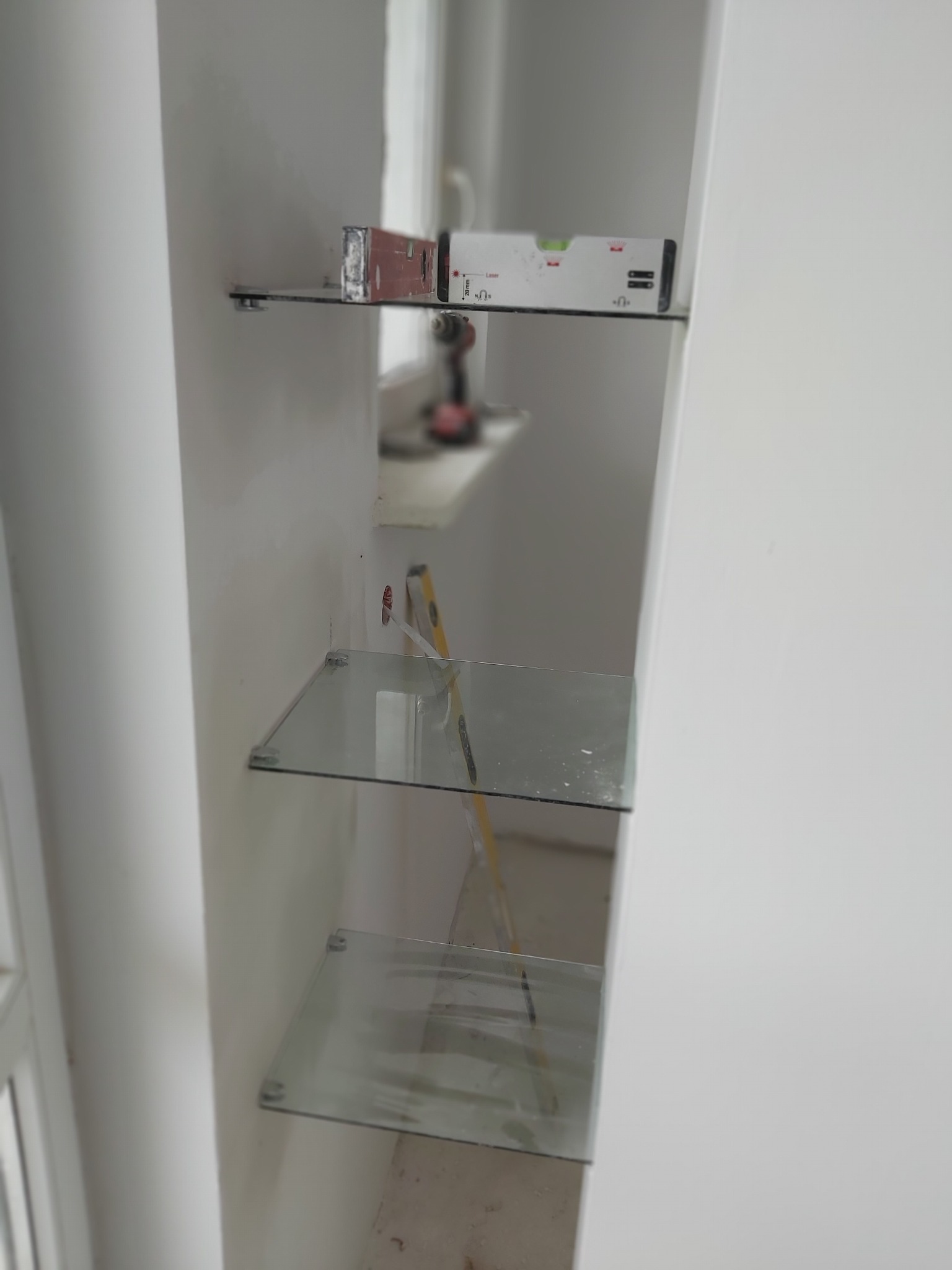 Montaż szklanych półek w wąskiej wnęce, widoczny poziomica, wiertarka i drabina oparta o ścianę