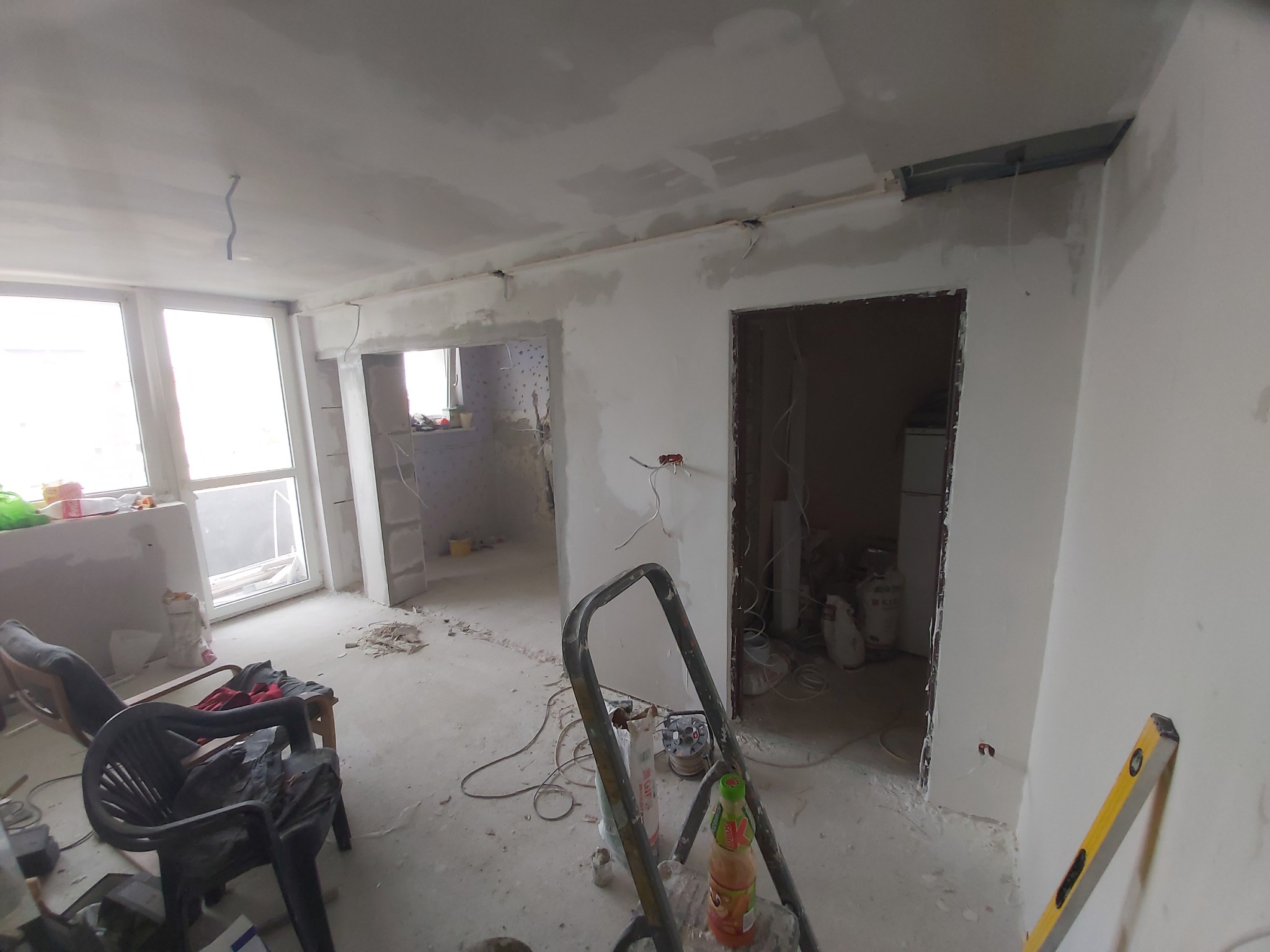 Wnętrze mieszkania w trakcie prac wykończeniowych; surowe ściany, widoczne ślady po szpachlowaniu, okablowanie wystające ze ściany, drabina malarska i narzędzia na podłodze, dwa krzesła w rogu pokoju.