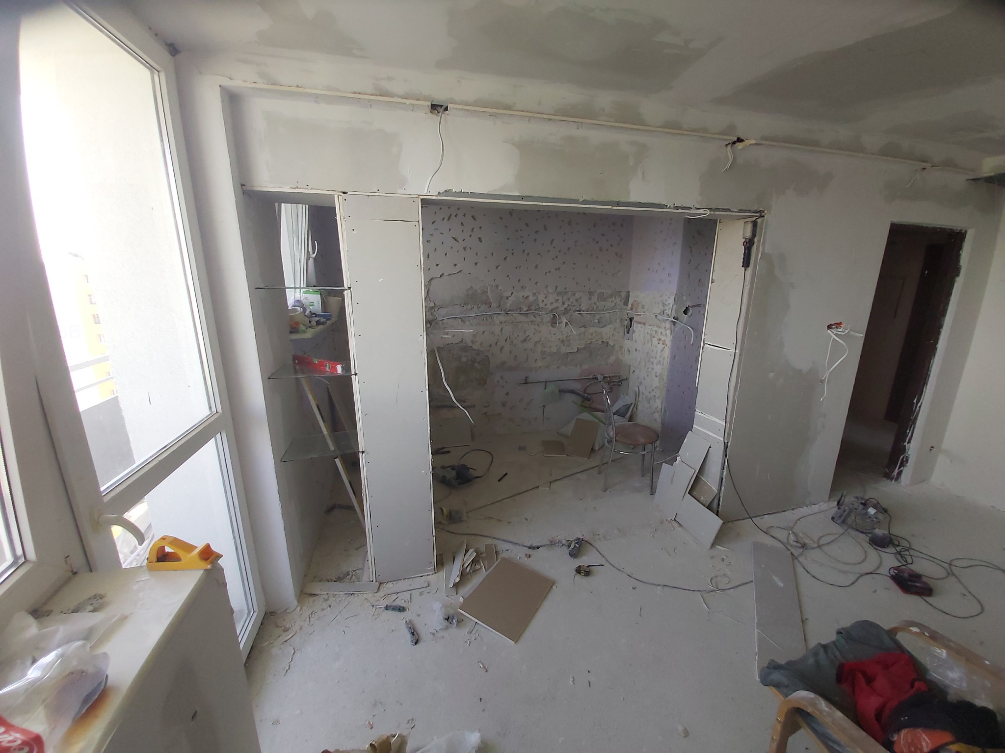 Wnętrze mieszkania w trakcie gruntownego remontu, widoczne ściany z odsłoniętą instalacją elektryczną, częściowo zabudowane płyty gipsowo-kartonowe, narzędzia i materiały budowlane rozrzucone...