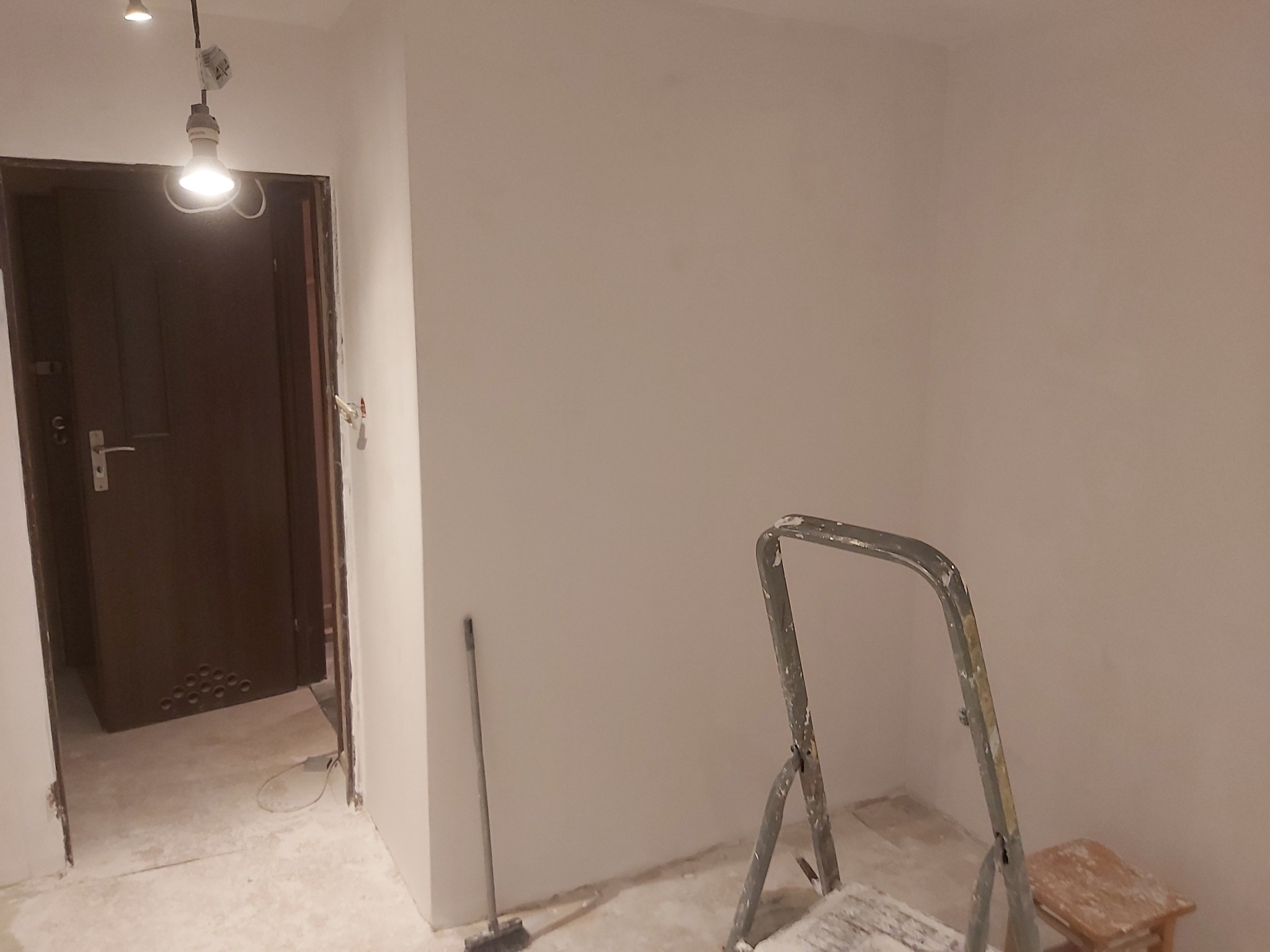 Świeżo pomalowane wnętrze z widoczną drabiną malarską, niedoskonałościami na podłodze i otwartymi drzwiami z widocznymi otworami wentylacyjnymi w dolnej części, oświetlone pojedynczą żarówką