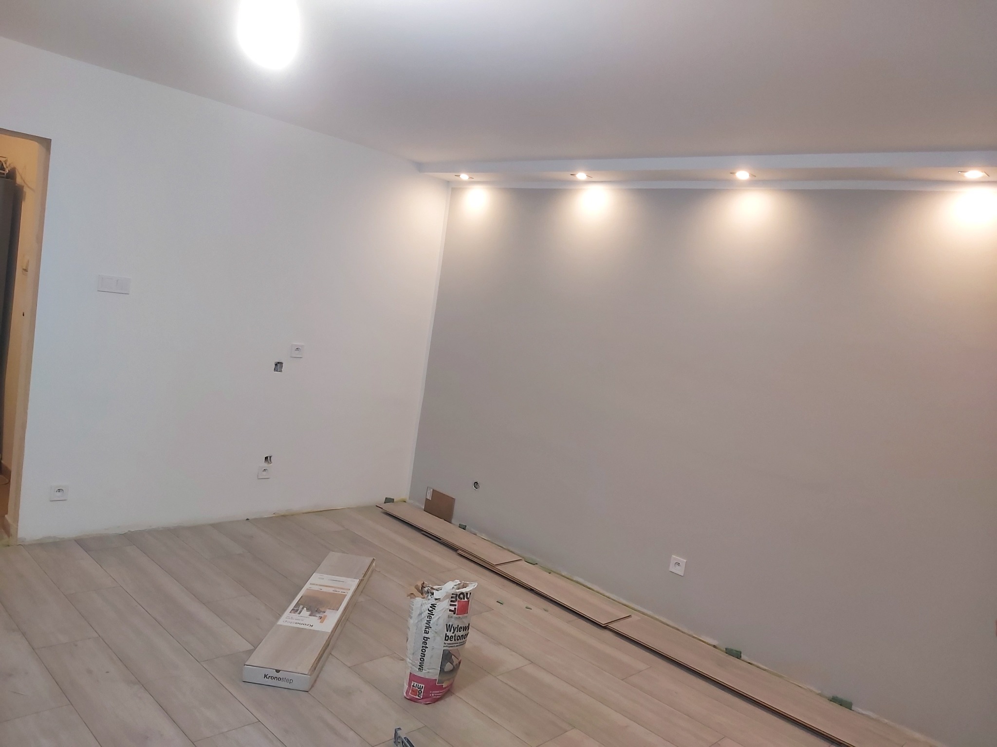 Wnętrze pokoju w trakcie układania jasnych paneli podłogowych, widoczny pakiet paneli Kronostep oraz worek z wylewką betonową BauMit, listwy przypodłogowe jeszcze nie zamontowane.