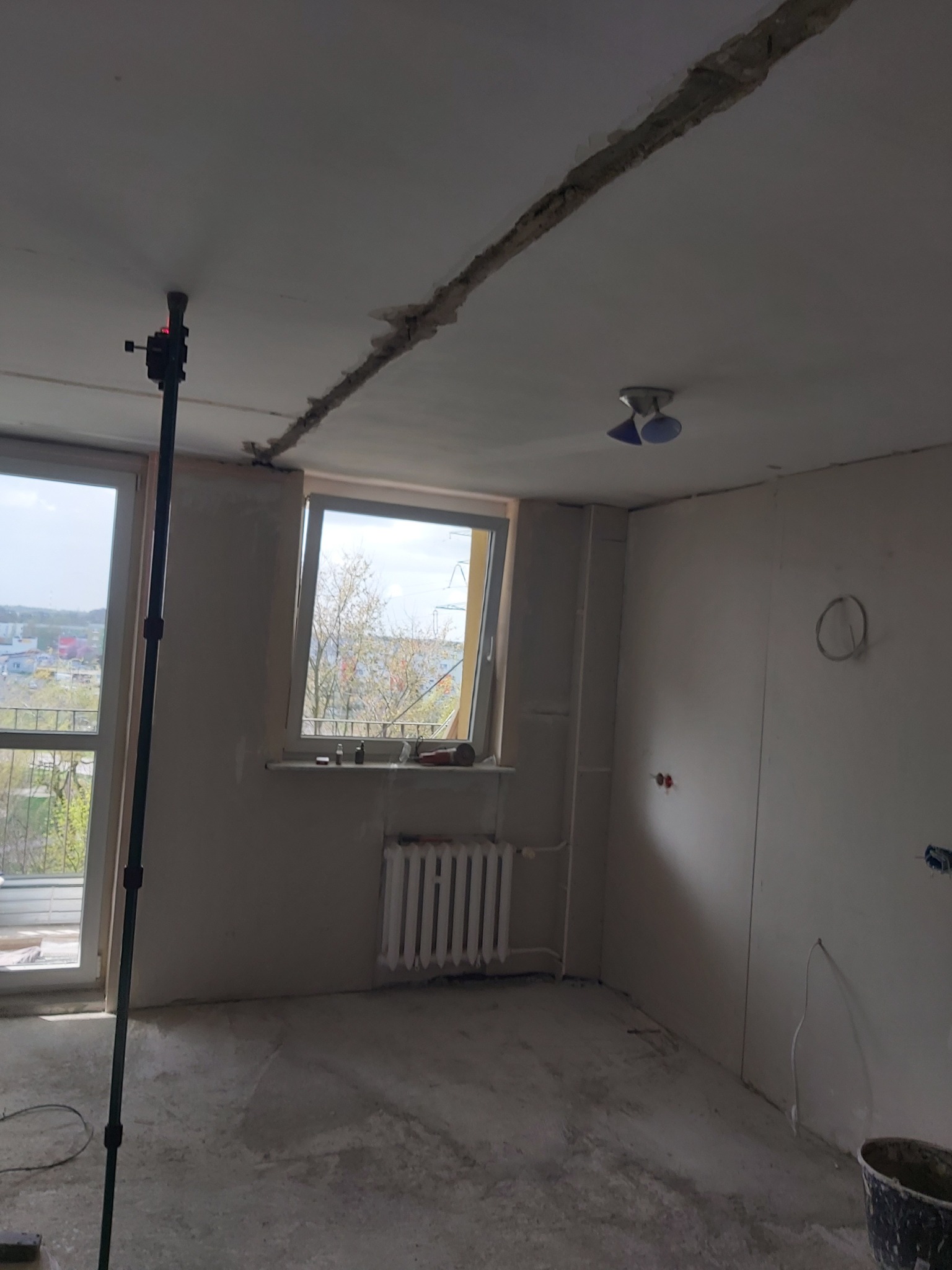 Wnętrze mieszkania w trakcie prac remontowych: odsłonięta instalacja elektryczna w suficie, widoczne ślady po kuciu, grzejnik, okno z widokiem na miasto, poziomica laserowa na statywie.