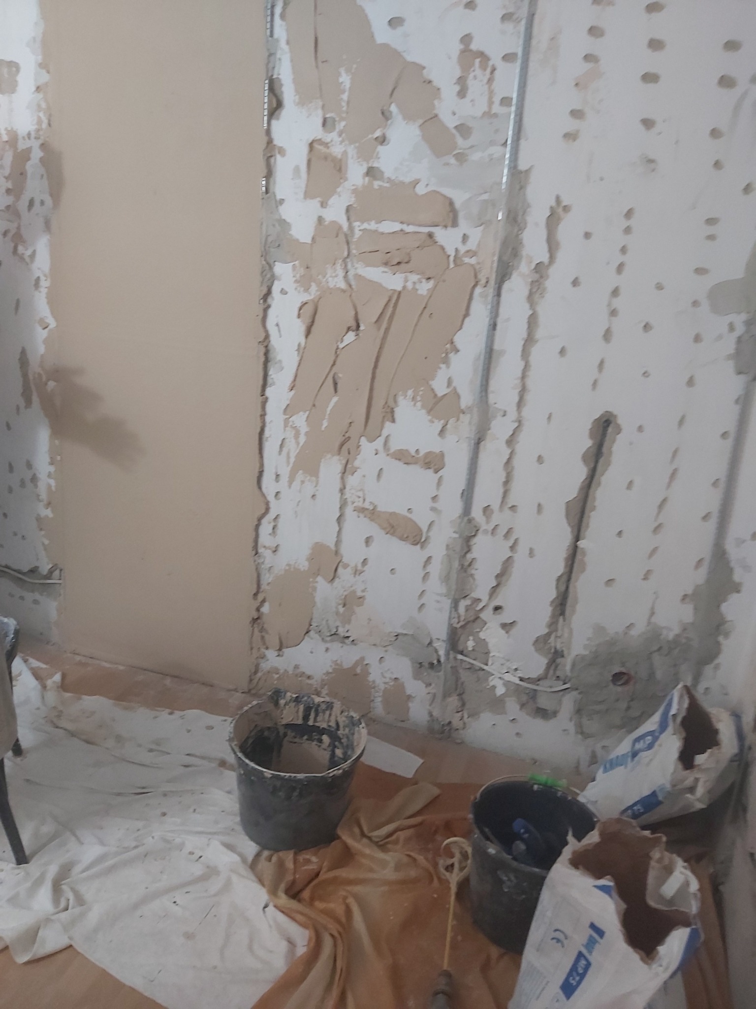 Ściana w trakcie remontu: częściowo otynkowana, z widocznymi śladami po starym tynku, metalowe profile, wiadro i worki z zaprawą na podłodze zabezpieczonej folią malarską.