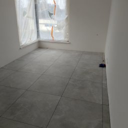 MarkBau - Pokój po ułożeniu szarych, marmurkowych płytek podłogowych, okno zabezpieczone folią malarską, widoczne drobne zabrudzenia po remoncie.