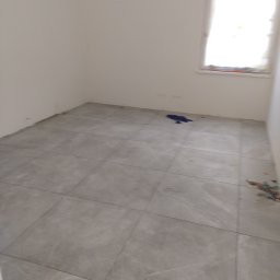 MarkBau - Ułożone szare płytki podłogowe w pustym pokoju z oknem, widoczne niebieskie szmaty i pozostałości po montażu płytek przy ścianach.