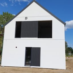 MarkBau - Nowoczesny, dwukondygnacyjny dom w trakcie budowy, z białymi ścianami i grafitowymi detalami, widoczne otwory okienne i przesuwne panele, niezagospodarowany teren wokół.