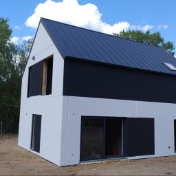 MarkBau - Nowoczesny dom jednorodzinny w trakcie wykończenia elewacji, z białą fasadą, czarnym dachem i elementami wykończeniowymi, widoczne niezabudowane otwory okienne i drzwiowe, otoczenie placu budowy.