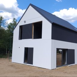 MarkBau - Nowoczesny dom dwukondygnacyjny z białą elewacją i ciemnoszarą górną częścią oraz dachem z blachy, w trakcie wykańczania, z widocznymi oknami i drzwiami, na tle nieba z chmurami i drugiego domu...