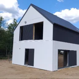 Nowoczesny dom dwukondygnacyjny z białą elewacją i ciemnoszarą górną częścią oraz dachem z blachy, w trakcie wykańczania, z widocznymi oknami i drzwiami, na tle nieba z chmurami i drugiego domu...
