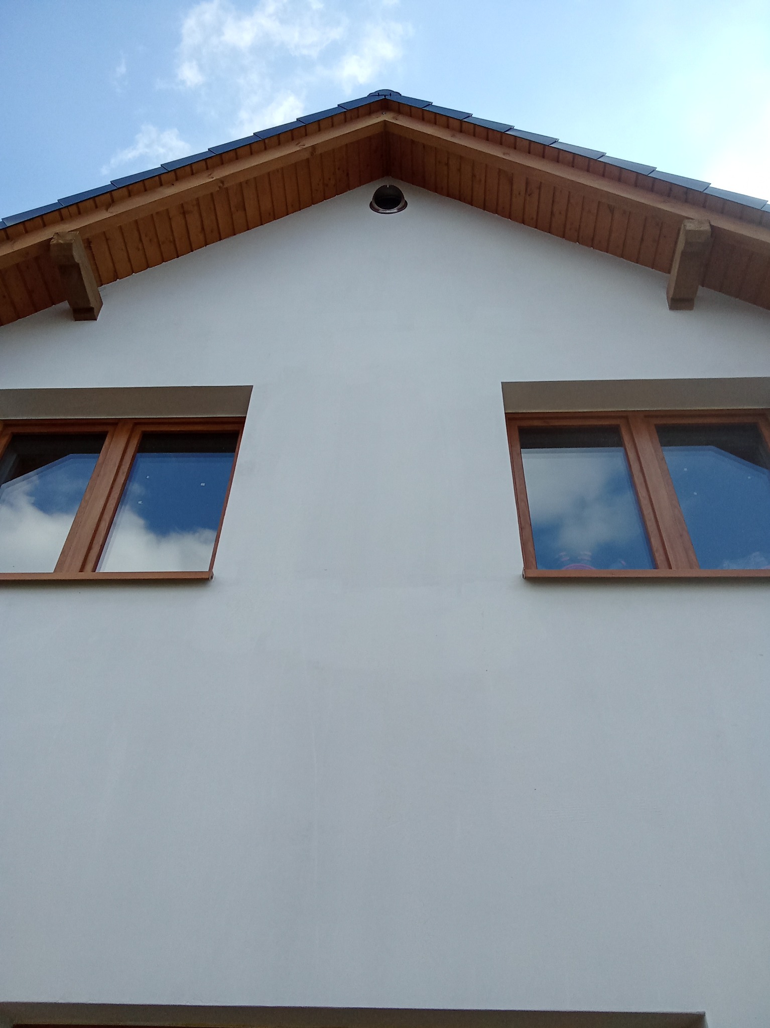 Elewacja domu jednorodzinnego w Skorzewie po tynkowaniu, widoczne dwa okna z drewnianą ramą i fragment dachu z drewnianą podbitką.