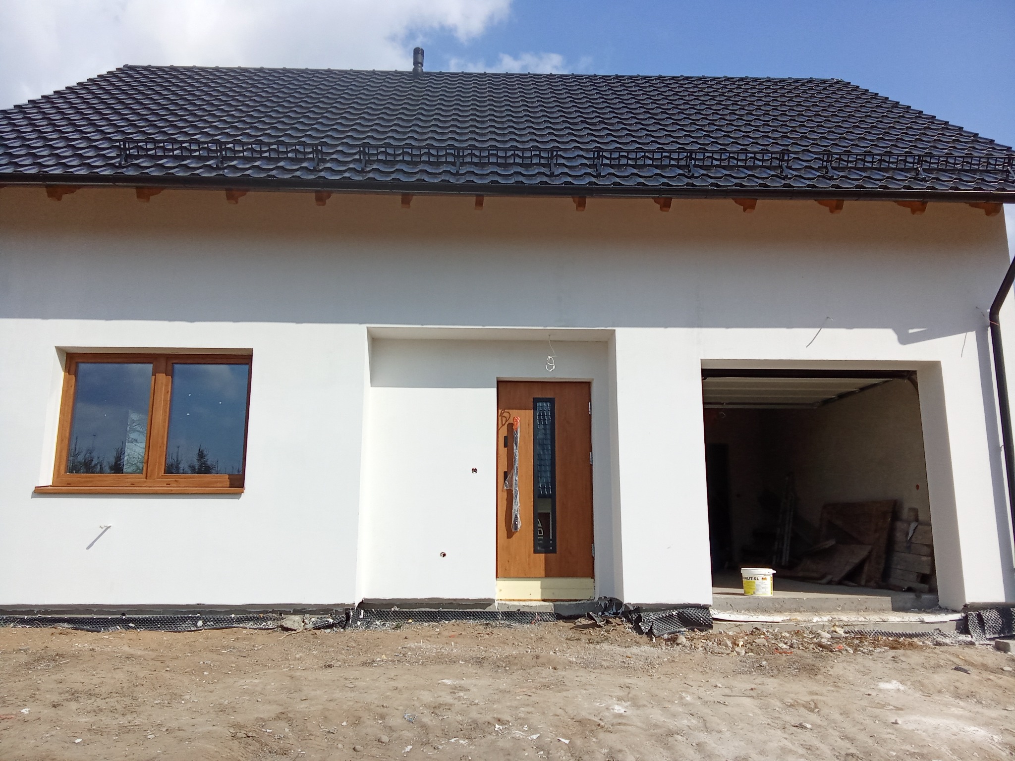 Nowo wybudowany dom jednorodzinny z ciemną dachówką, drewnianymi oknami i drzwiami wejściowymi; widoczne ślady prac wykończeniowych wokół budynku.