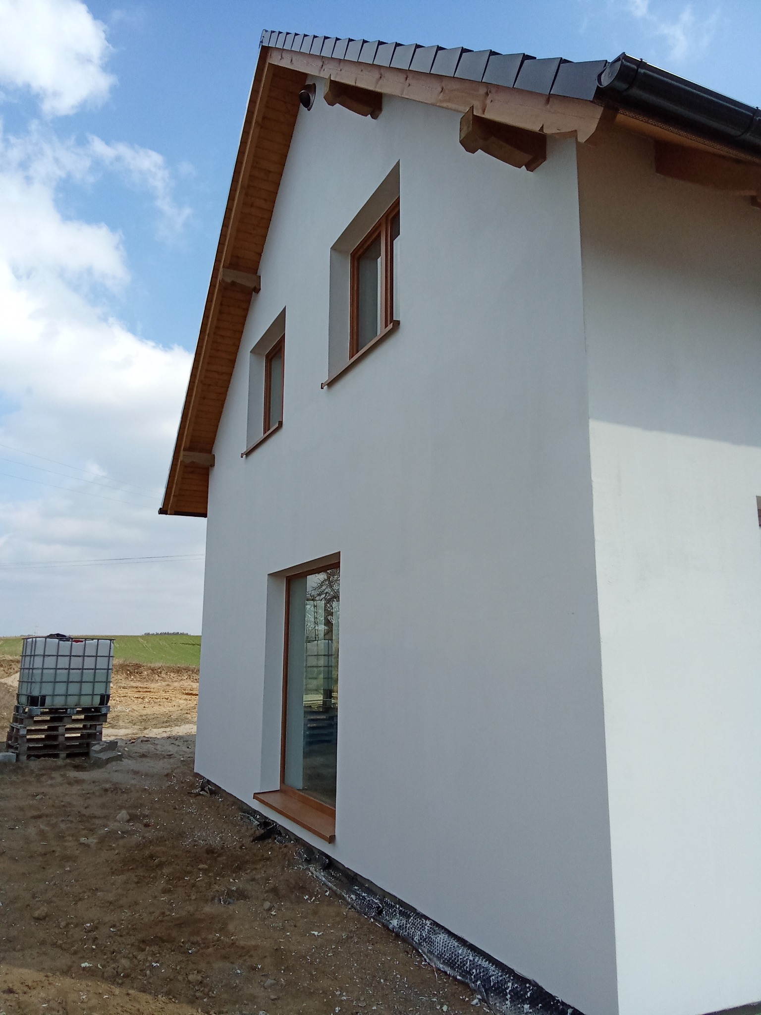 Boczny widok nowo otynkowanego domu jednorodzinnego z drewnianą stolarką okienną i dachem z ciemnej dachówki, na tle błękitnego nieba z chmurami, z widocznym zbiornikiem IBC na palecie w tle.