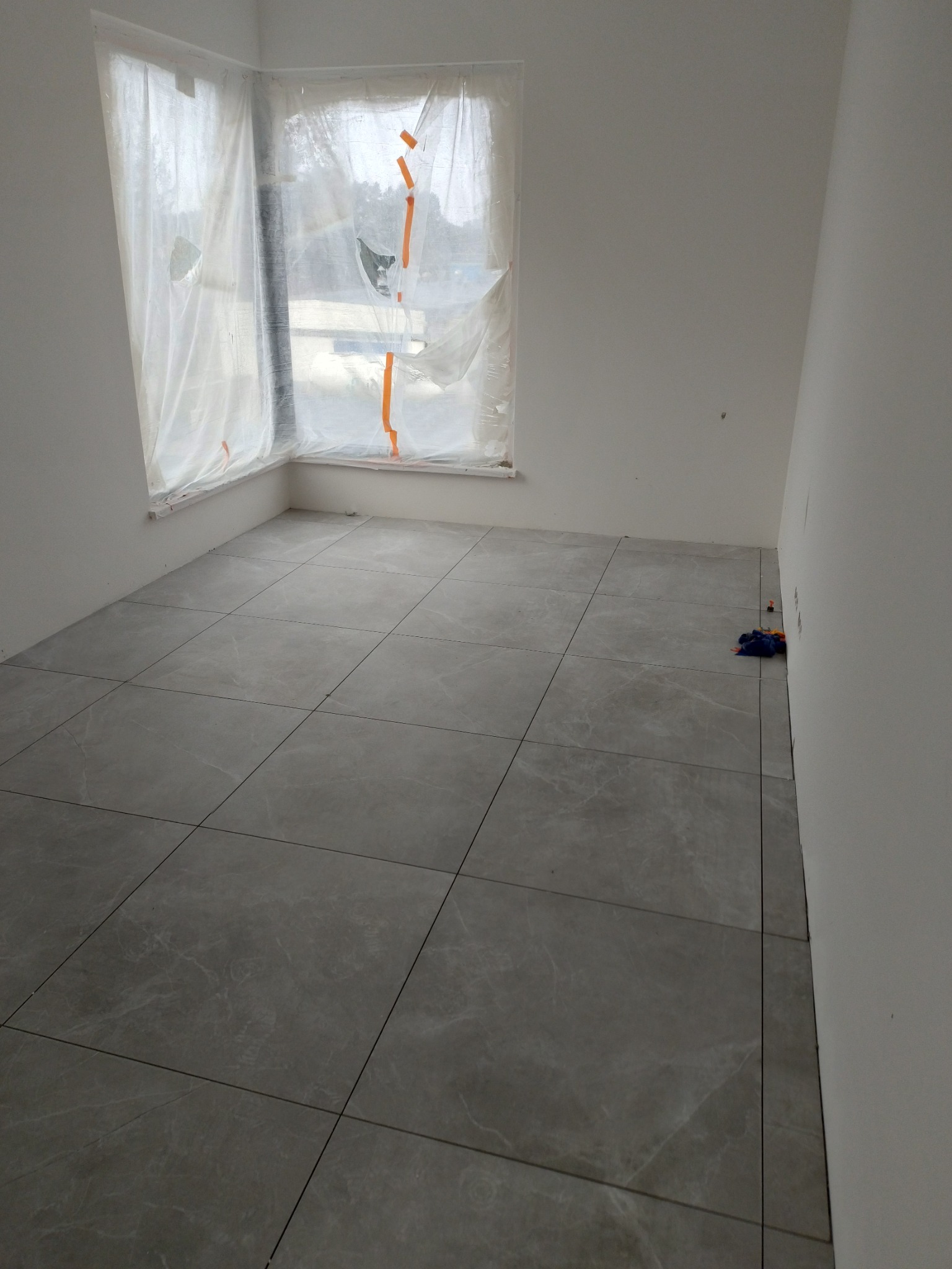 Pokój po ułożeniu szarych, marmurkowych płytek podłogowych, okno zabezpieczone folią malarską, widoczne drobne zabrudzenia po remoncie.