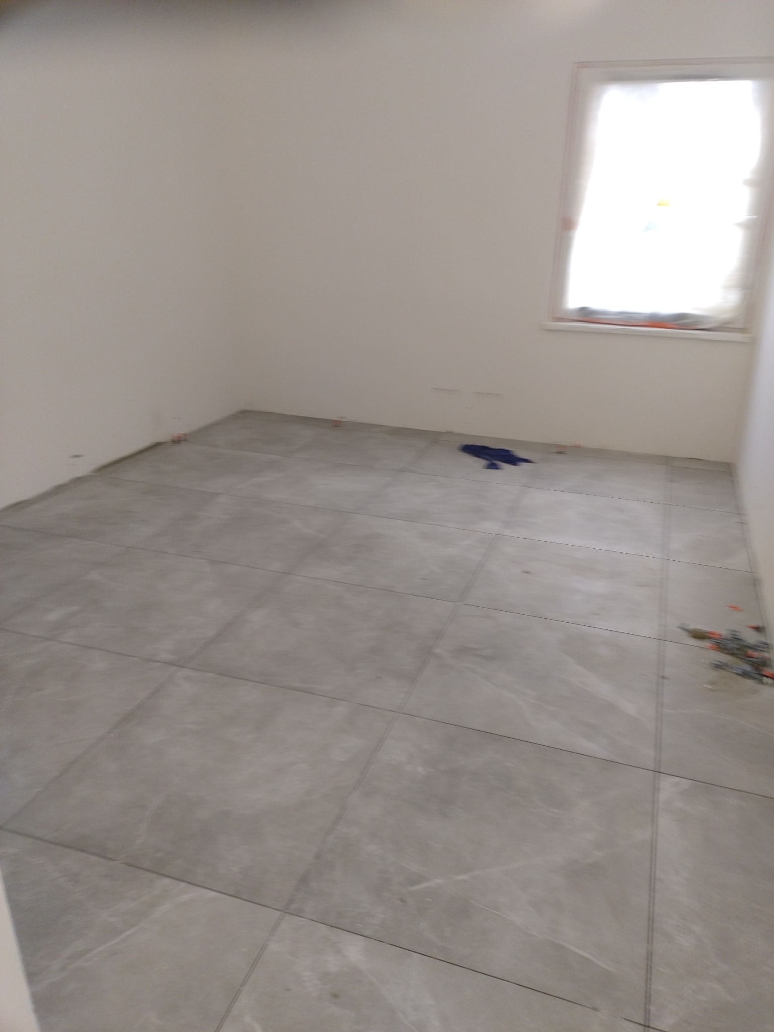 Ułożone szare płytki podłogowe w pustym pokoju z oknem, widoczne niebieskie szmaty i pozostałości po montażu płytek przy ścianach.