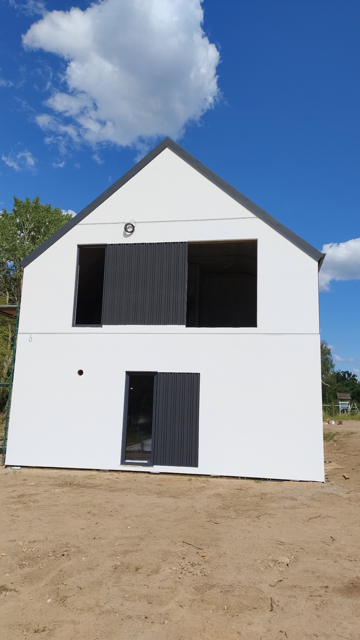 Nowoczesny, dwukondygnacyjny dom w trakcie budowy, z białymi ścianami i grafitowymi detalami, widoczne otwory okienne i przesuwne panele, niezagospodarowany teren wokół.