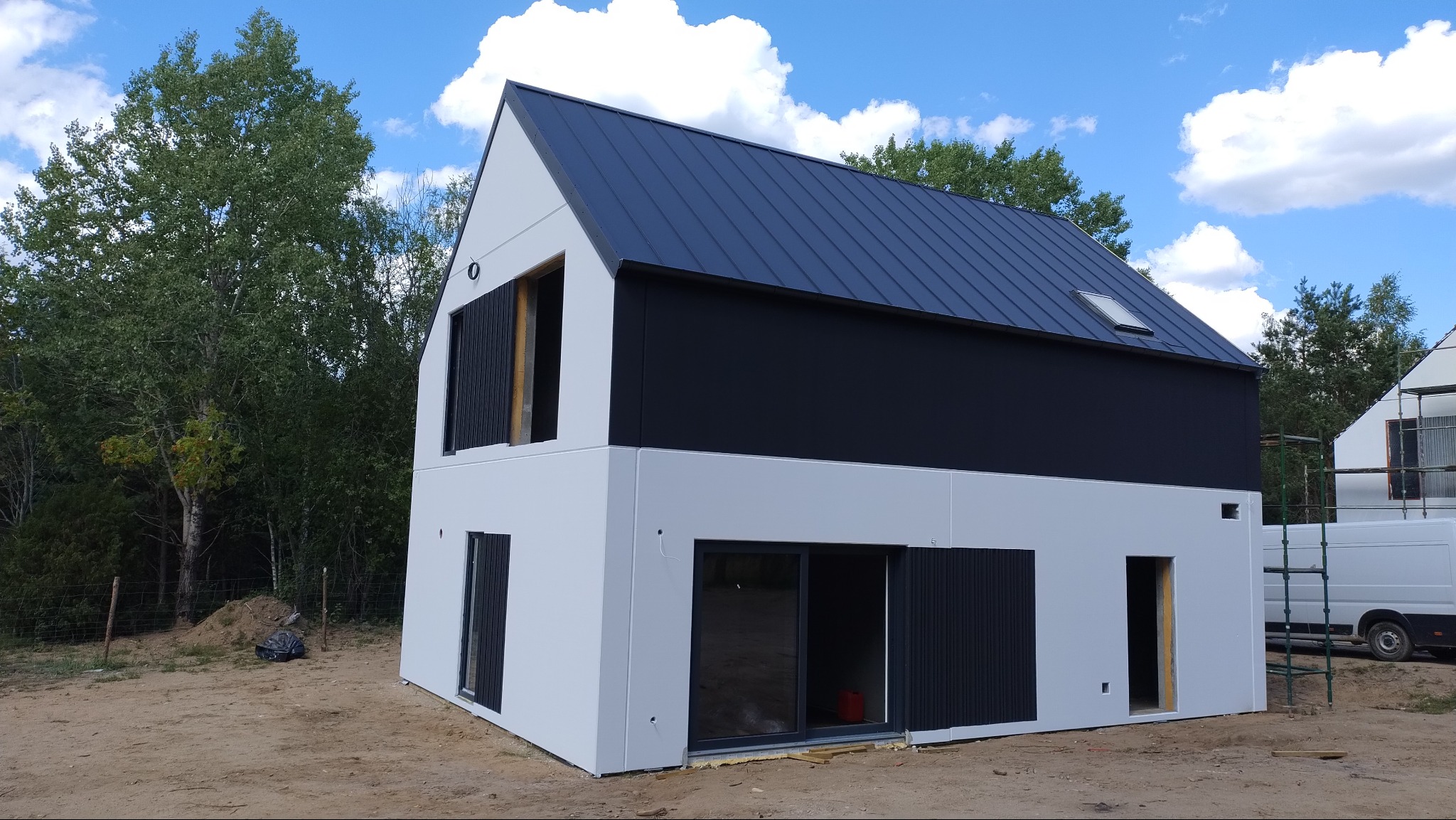 Nowoczesny dom jednorodzinny w trakcie wykończenia elewacji, z białą fasadą, czarnym dachem i elementami wykończeniowymi, widoczne niezabudowane otwory okienne i drzwiowe, otoczenie placu budowy.