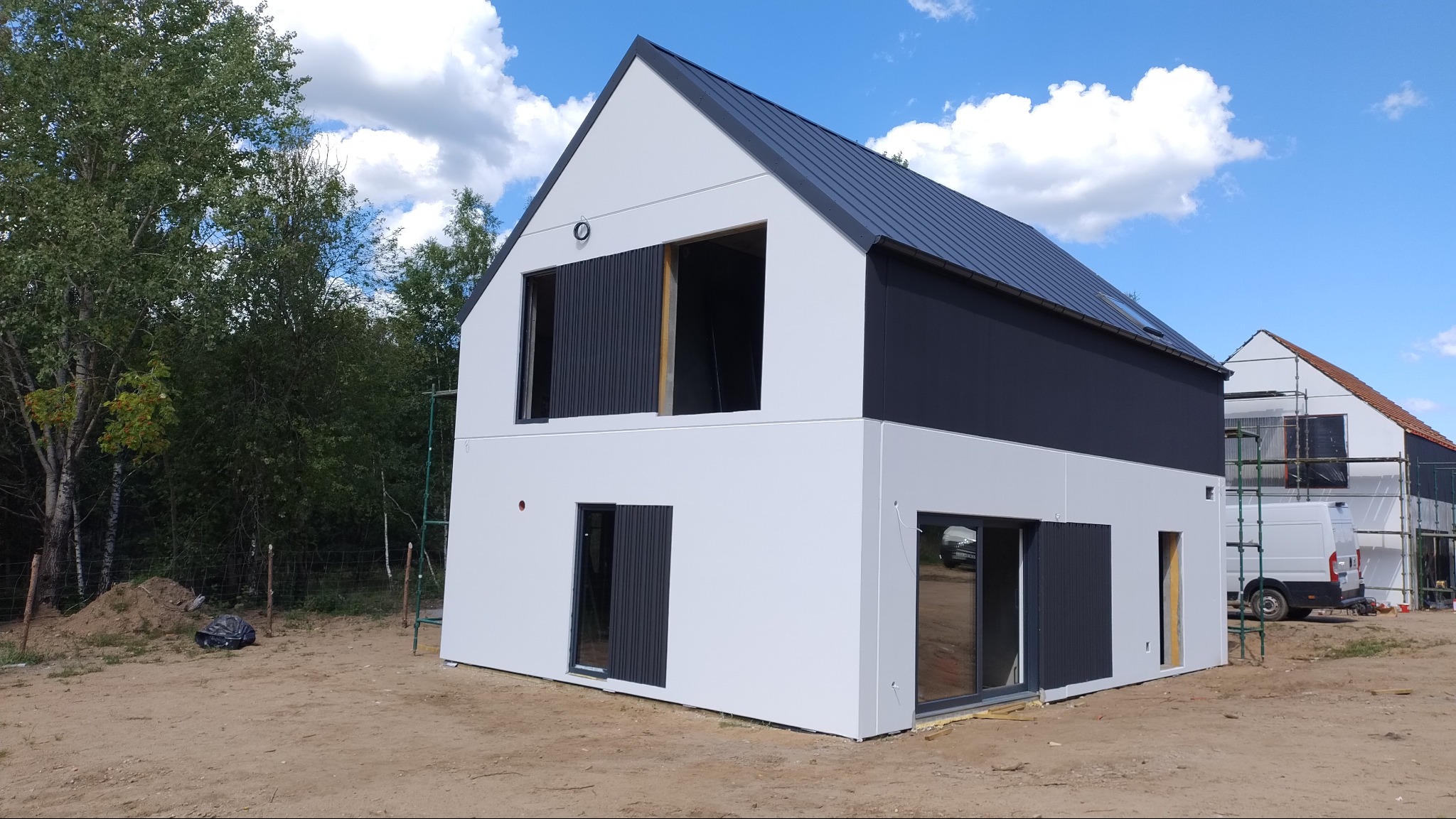 Nowoczesny dom dwukondygnacyjny z białą elewacją i ciemnoszarą górną częścią oraz dachem z blachy, w trakcie wykańczania, z widocznymi oknami i drzwiami, na tle nieba z chmurami i drugiego domu...