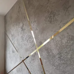 Dekoracyjna ściana z szarego betonu architektonicznego z wstawkami z błyszczących, złotych listew tworzących geometryczny wzór.