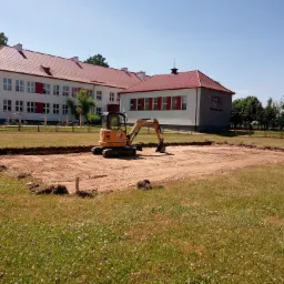 Mini koparka CAT 302.7D na terenie budowy w Szyszkach, przygotowuje wykop pod fundament budynku szkolnego z czerwonym dachem.