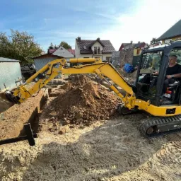 Żółta mini koparka w trakcie wykonywania wykopu pod fundamenty budynku, operator w kabinie, widoczne ślady gąsienic na piaszczystym podłożu, w tle zabudowa jednorodzinna.
