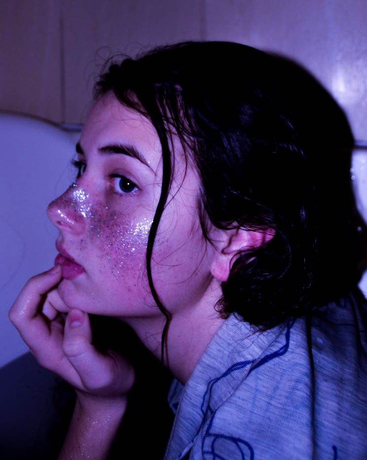 Portret kobiety z brokatem na twarzy, mokre włosy, ujęcie z profilu, pastelowe oświetlenie.