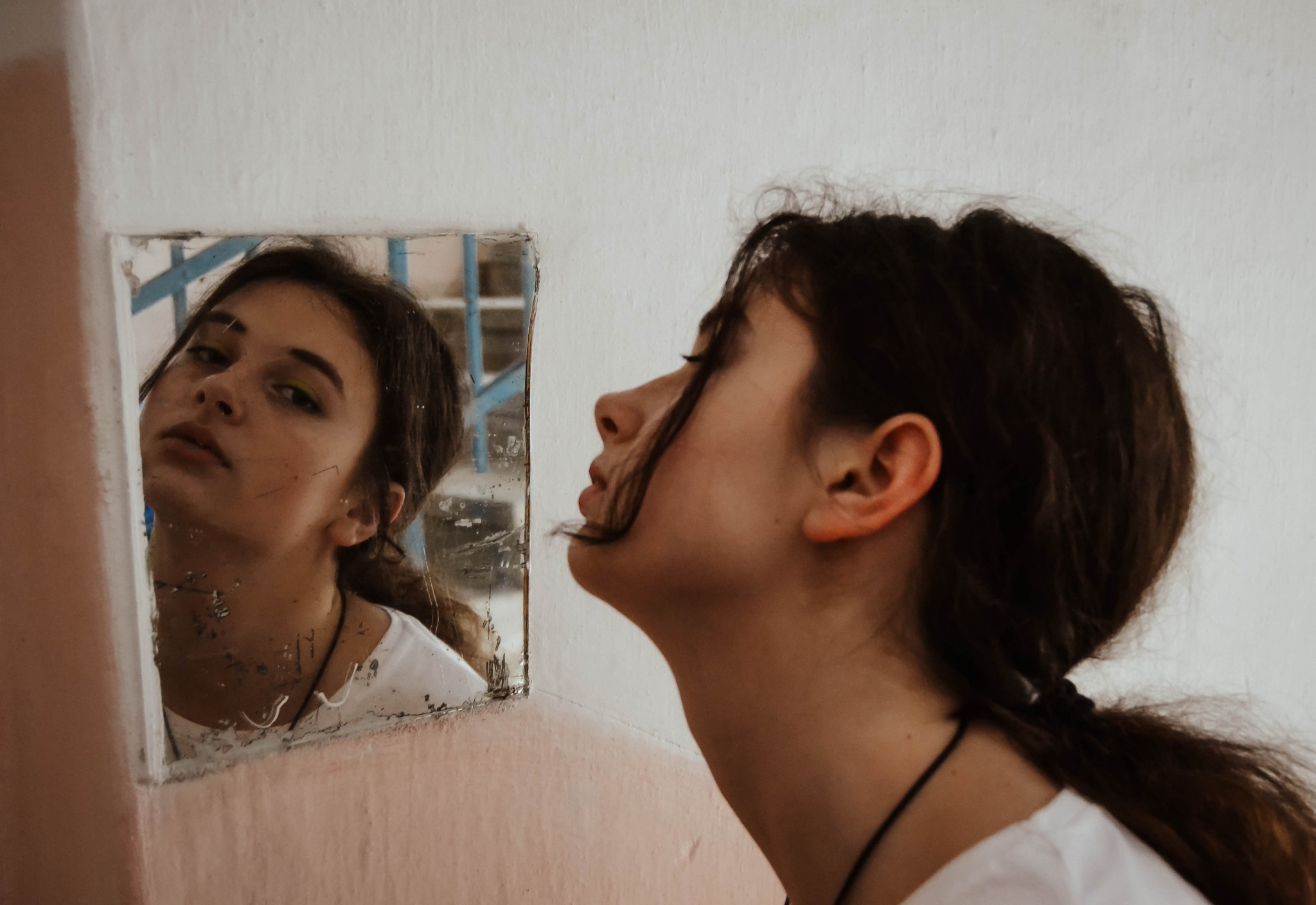 Portret młodej kobiety spoglądającej w małe, brudne lustro przy ścianie, widoczny profil szyi i odbicie twarzy z lekkim makijażem oczu.
