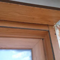 Fragment drewnianej ramy okiennej z widocznym fragmentem szyby i nieotynkowanym murem z cegły obok, ukazujący etap montażu.