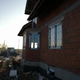 Surowy stan budynku w trakcie montażu okien z białymi ramami, widoczne cegły, niezabezpieczone fundamenty i gruz wokół.