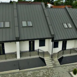 Nowoczesne segmenty bliźniacze z szarym dachem z blachy na rąbek stojący, czarnymi oknami, drzwiami i balustradami, oraz małymi oknami dachowymi.