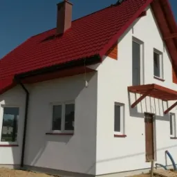 Nowo wybudowany, biały dom jednorodzinny z czerwoną dachówką, widoczne okna z białymi ramami, brązowe drzwi wejściowe z drewnianym zadaszeniem, teren wokół domu w trakcie zagospodarowywania.