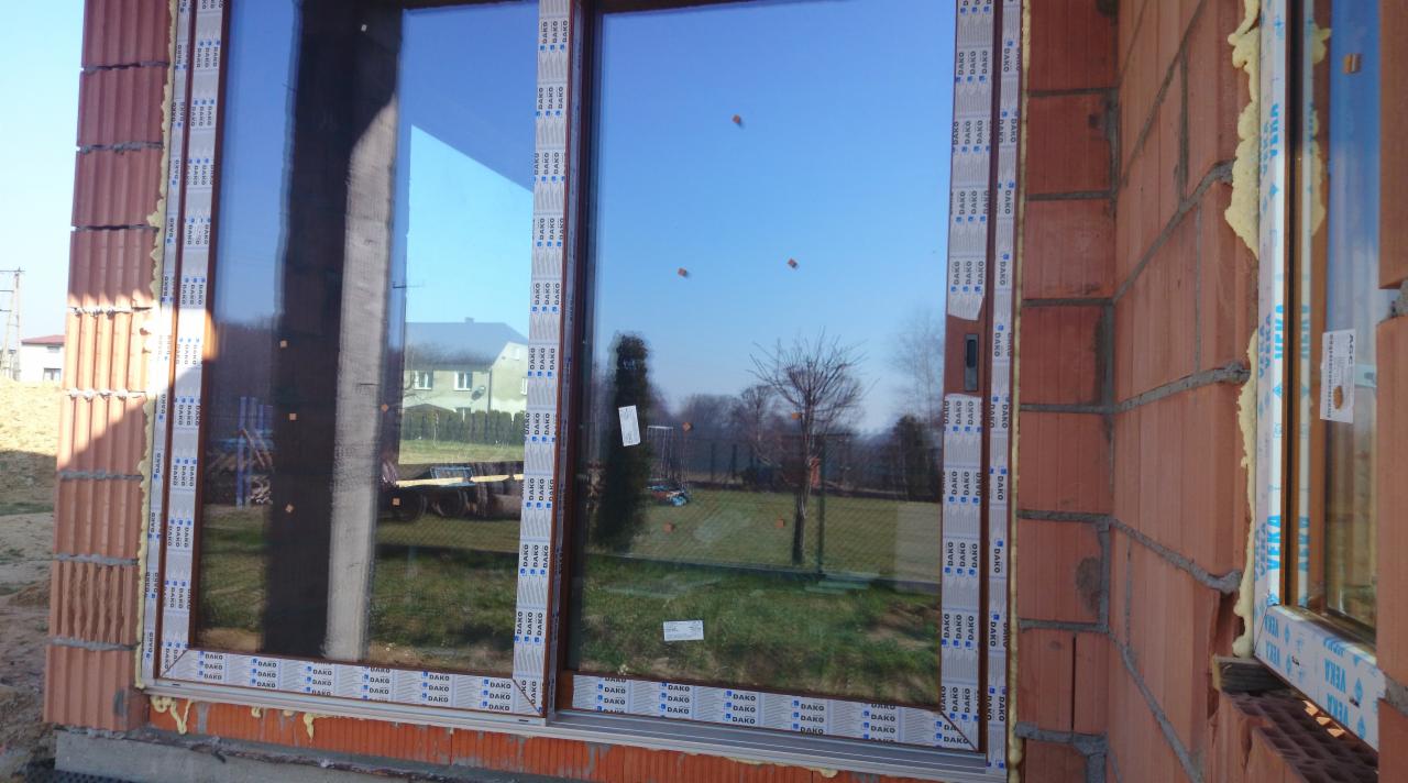 Montaż okna w surowym stanie, widoczne cegły i pianka montażowa, oklejone profile z logo producenta, w odbiciu szyby krajobraz z domem.