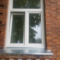 Białe okno PCV zamontowane w murze z czerwonej cegły, z widocznym metalowym parapetem zewnętrznym.