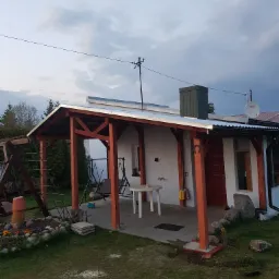 Drewniana wiata przydomowa z pomalowanymi na pomarańczowo słupami, widok z zewnątrz na ogród i dom z elementami dekoracyjnymi, takimi jak podkowa nad drzwiami.