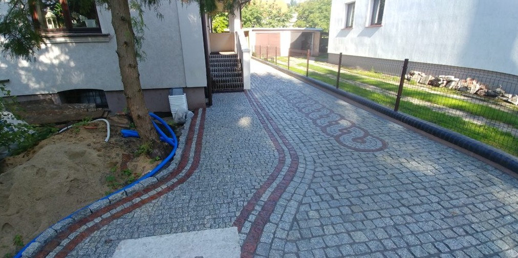 Ukończony podjazd z szarej kostki granitowej z bordiurą i ozdobnym wzorem w kształcie logo samochodowego, widok z góry na tle domu i ogrodu.