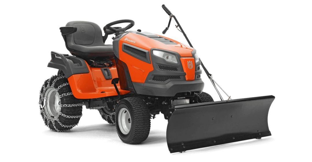 Pomarańczowy traktorek ogrodowy Husqvarna z łańcuchami na tylnych kołach i zamontowanym pługiem do śniegu, prezentowany na białym tle.