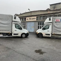 Dwa białe busy dostawcze, jeden z logo MGTrans, stojące naprzeciwko siebie na mokrym asfalcie w Sosnowcu, z budynkiem firmy Polbrass w tle.